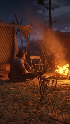 a man sitting in front of a fire next to a tent on top of a field