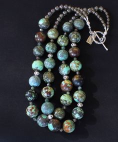 This striking Necklace showcases 2 strands of 20mm Turquoise Rounds. The stones are multi-hued, displaying varying hues of blue and green with brown & black matrix. The blend of color and pattern is eye-catching. The Turquoise is beautifully shaped and polished to a soft sheen. The Turquoise is paired with several complementary elements, including 6mm luminous green Czech Druk Glass Rounds, 6mm green Rutilated Quartz Rounds, 5mm Peridot Rounds, and 6mm transparent green Fire Polished Glass Ovals Turquoise Double Strand Gemstone Jewelry, Turquoise Multi-stone Round Necklace, Fusion Style Turquoise Round Bead Jewelry, Fusion Style Turquoise Round Beads Jewelry, Artisan Double Strand Turquoise Jewelry, Multicolor Chrysocolla Round Beads Jewelry, Fusion Style Turquoise Jewelry With Gemstone Beads, Turquoise Fusion Jewelry With Gemstone Beads, Hues Of Blue