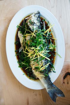 a white plate topped with fish covered in sauce and greens on top of a wooden table