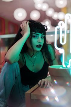 a woman with black makeup sitting in front of a neon sign and looking at the camera