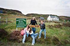 three women standing in front of a sign on top of a grass covered hill with houses behind them