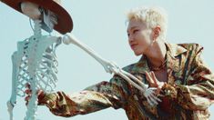 a woman with white hair is holding a skeleton and wearing a hat while standing in front of a blue sky