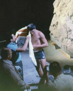 a man in swim trunks standing next to another man with a towel around his neck