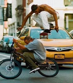 two men riding bikes in the street with cars behind them and one man doing tricks on his bike