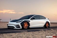 a white car parked on top of a sandy beach next to the ocean at sunset