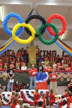 a large group of people standing in front of an olympic sign with balloons hanging from the ceiling