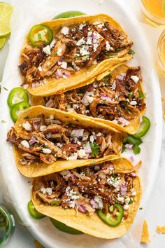 three tacos with shredded meat, onions and peppers on a white platter next to limes