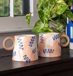 two coffee mugs sitting on top of a table next to a potted plant