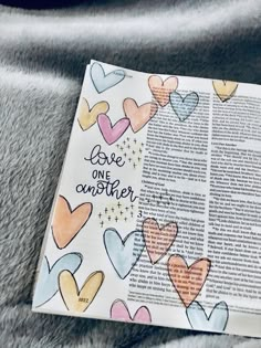 an open bible with hearts painted on it and the words love one another in rainbow colors