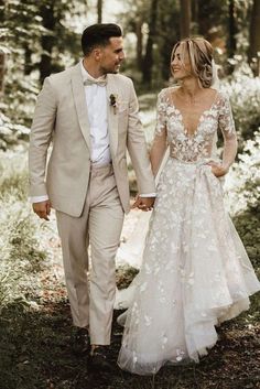 a bride and groom are walking through the woods together in their wedding attire, holding hands