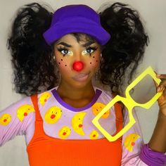 a woman with clown makeup holding up a pair of yellow glasses and wearing a purple hat