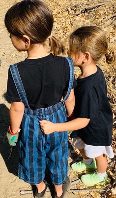 two young children standing next to each other on a dirt ground covered in leaves and grass