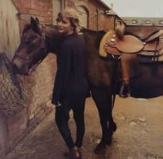 a woman standing next to a horse in front of a brick building with a saddle on it