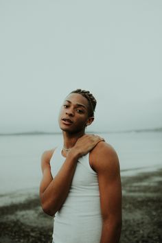 a man with dreadlocks standing by the water wearing a white tank top and looking off into the distance