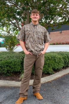 a man standing in front of a tree wearing brown pants and a camo shirt