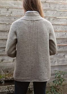 a woman standing in front of a wooden fence wearing a gray sweater and black jeans