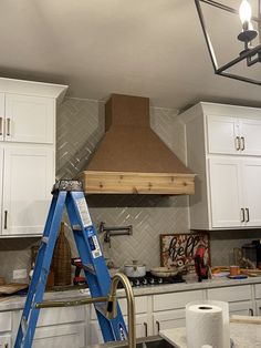 a kitchen with white cabinets and a blue ladder in front of a stove hood that is being installed