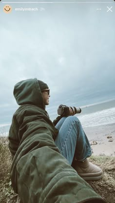 a man sitting on top of a grass covered field next to the ocean holding a camera