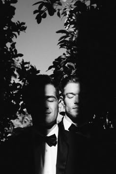 two men in tuxedos standing next to each other under a tree with their eyes closed