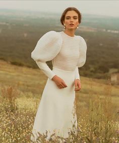 a woman in a white dress standing on top of a grass covered hill with her hands on her hips