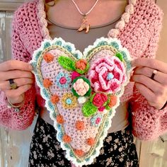 a woman is holding a crocheted heart with flowers on the front and sides