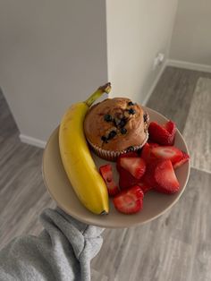 a person holding a plate with a muffin, strawberries and banana on it