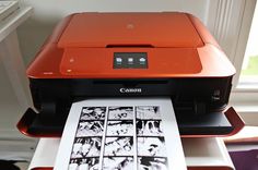 an orange printer sitting on top of a white table next to a window with pictures