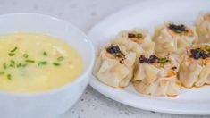 a white plate topped with dumplings next to a bowl filled with yellow liquid and garnished with chives