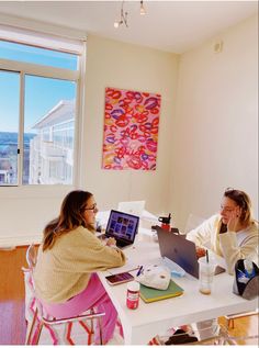 two women sitting at a table working on their laptops in front of a window