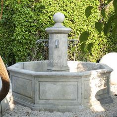 an outdoor fountain in the middle of a gravel area with trees and bushes behind it