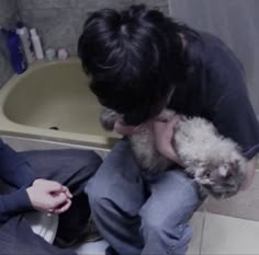 two people sitting on the floor in front of a bath tub holding a dog and petting it