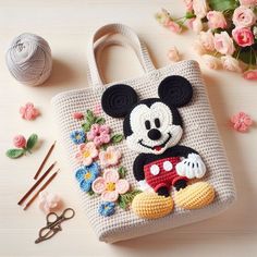 a crocheted bag with a mickey mouse on it next to flowers and knitting needles