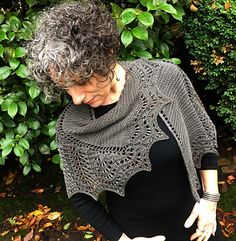 a woman standing in front of some bushes wearing a gray shawl and black shirt