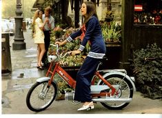 a woman riding on the back of a red bike in front of a flower shop