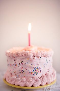 a pink birthday cake with sprinkles and a single lit candle on top