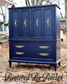 a blue armoire with gold handles in front of some trees and leaves on the ground