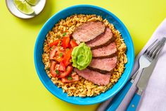 a blue bowl filled with meat and rice next to a fork, knife and spoon