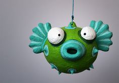 a green ornament with two eyes hanging from a string on a wall in front of a gray background