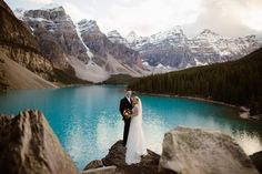 a bride and groom standing on the edge of a mountain lake