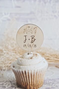 a cupcake with white frosting and a wooden name tag on top that says j b