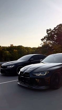 two black cars driving down the road next to each other in front of some trees