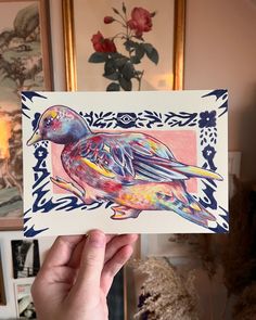 a hand holding up a card with a bird on it's side and flowers in the background