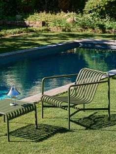 two chairs and a table in the grass near a pool with a lamp on it