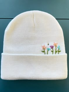 a white beanie with flowers embroidered on the front and side, sitting against a blue background