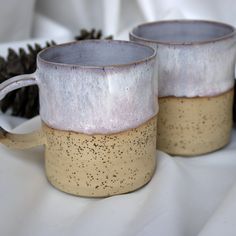 two coffee mugs sitting next to each other on a white tablecloth with pine cones in the background