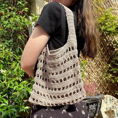 a woman carrying a crocheted purse on her back in front of some bushes