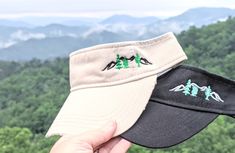 a person holding up two hats in front of some trees and mountains with green leaves on them