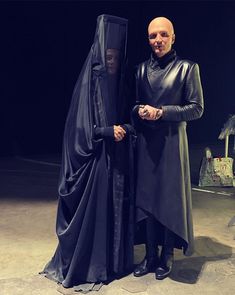 a man and woman dressed in black standing next to each other with one wearing a cape