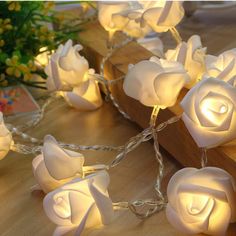 some white roses are lit up on a wooden table with string lights around the edges