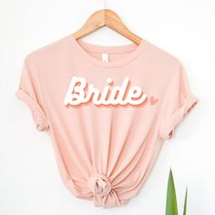 Share your excitement with this Bride Shirt. Perfect for brides-to-be! Whether your loved one is getting married or celebrating her wedding day, this shirt is sure to bring a smile. It's the perfect gift for all occasions: bachelorette parties, bridal showers, and weddings. ♥ Thank you for shopping with us! ♥ Please reach out with any questions or requests! :) How to Order: - Choose shirt color from the drop-down menu - Choose shirt size - Add to cart - If purchasing more than one item you will have to add it individually. One shirt is included in the price. Material: - Solid colors are 100% combed and ring-spun cotton - Heather colors are 52% combed and ring-spot cotton, 48% polyester - Ash colors are 99% combed and ring-spun cotton, 1% polyester - Fabric is pre-shrunk - Tear-away label - Pink Short Sleeve T-shirt For Bridal Shower, Pink Crew Neck Top For Bridal Shower, Pink Crew Neck Top For Wedding, Custom Text Short Sleeve Tops For Wedding, White Short Sleeve Top For Bridesmaids, White Custom Text Top For Wedding, White Custom Text T-shirt For Weddings, Pink Short Sleeve Top For Wedding, White Short Sleeve Bride T-shirt